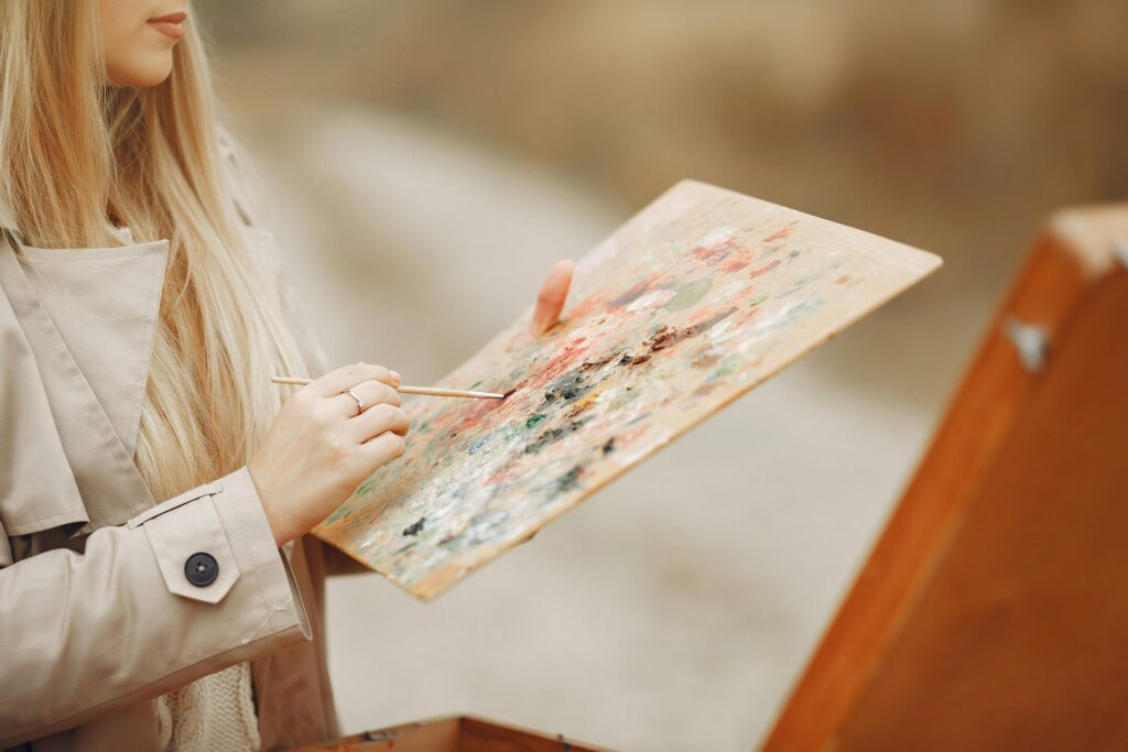 pexels-photo-4241308-4241308 Crop unrecognizable concentrated young female artist in trench coat using paintbrush and mixing colors on palette against wooden easel